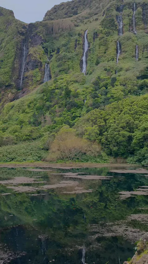 Dozens of thin waterfalls falling down a green cliff face into a still lagoon, Azores