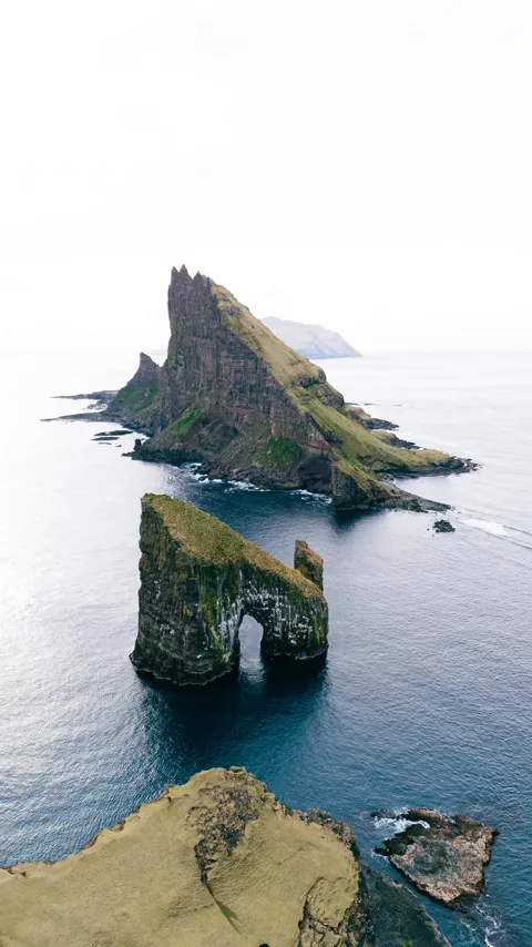 A sea stack with a natural arch standing off a green headland, Faroe Islands