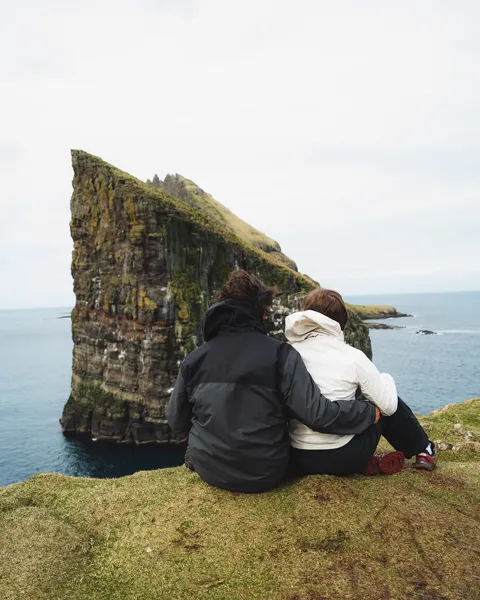 Two people sitting on a grassy edge looking across towering layered sea cliffs, Faroe Islands