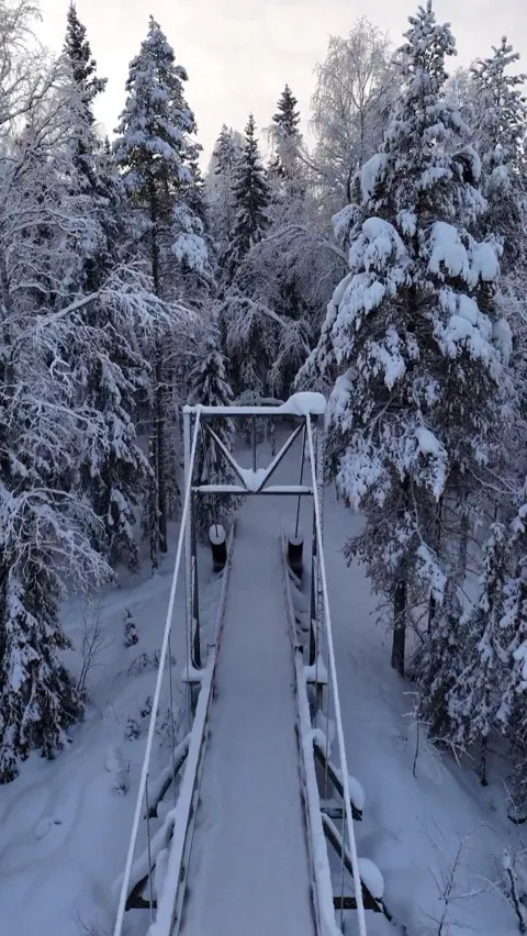 A footbridge crossing a frozen river through deep snow-covered forest, Lapland, Finland