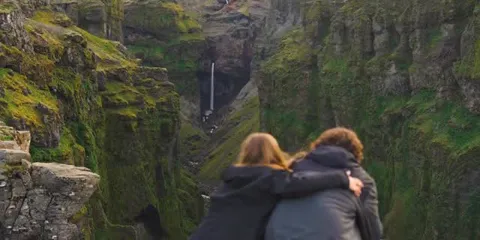 Two people at a viewpoint above a green canyon with a waterfall running through it, Iceland