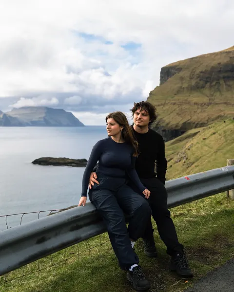 Kristi Leka sitting on a roadside guardrail above the sea with a green headland behind
