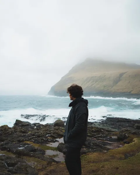 Kristi Leka on a rocky shoreline as cloud rolls over the headland behind, Faroe Islands